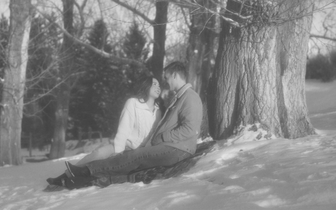 A couple sitting in the snow under bare winter trees.