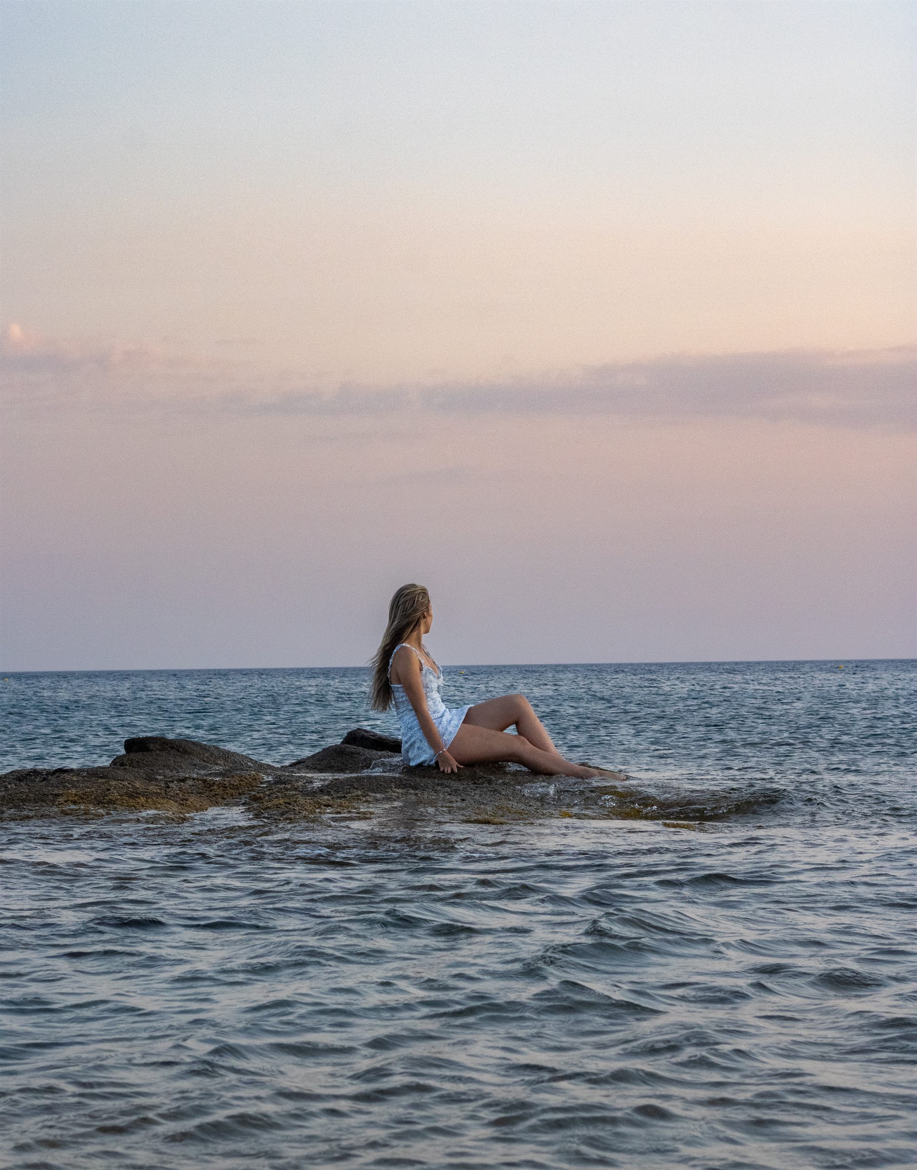 A woman seated on rocks at the water's edge under a soft pastel sky.