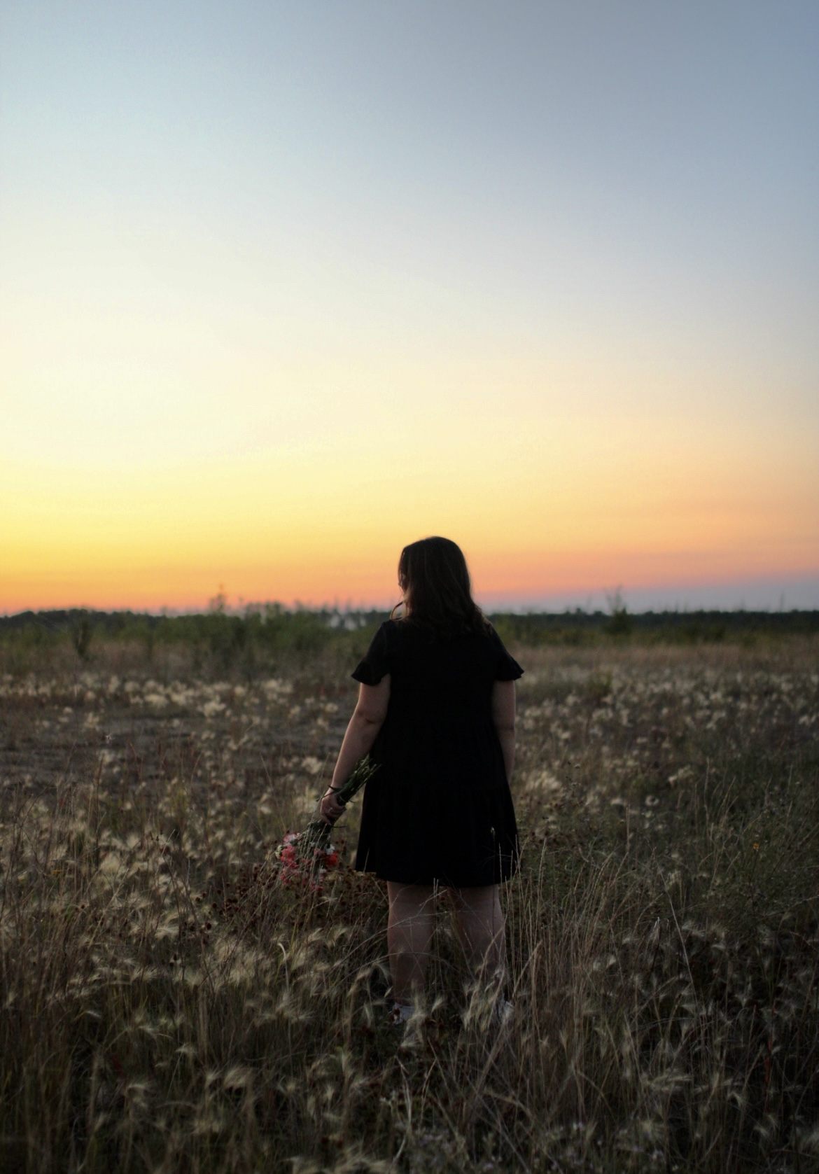A woman in a black dress holding wildflowers at sunset.