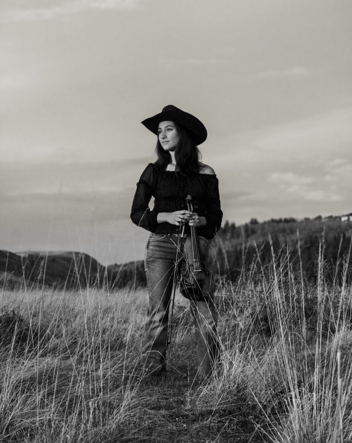 Black and white portrait of a woman in a hat holding a violin in tall grass.
